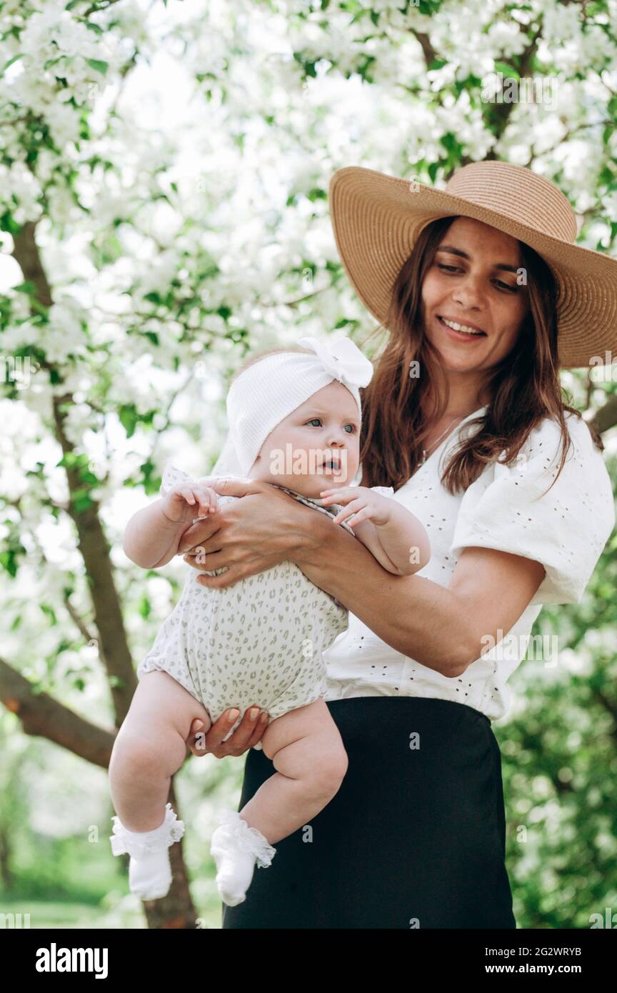 A cute beautiful young happy woman in a white shirt and straw hat is holding her smiling six month old daughter. Happy motherhood and happy childhood. Stock Photo