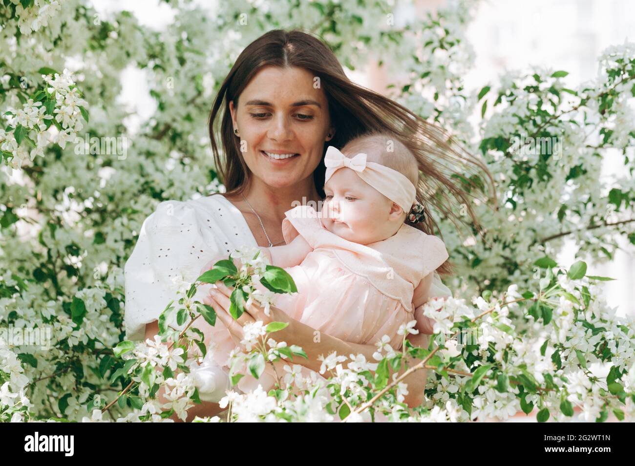 Portret cute beautiful young happy mom in a white shirt is holding her six month old daughter in a pink dress. Happy motherhood and happy childhood. P Stock Photo
