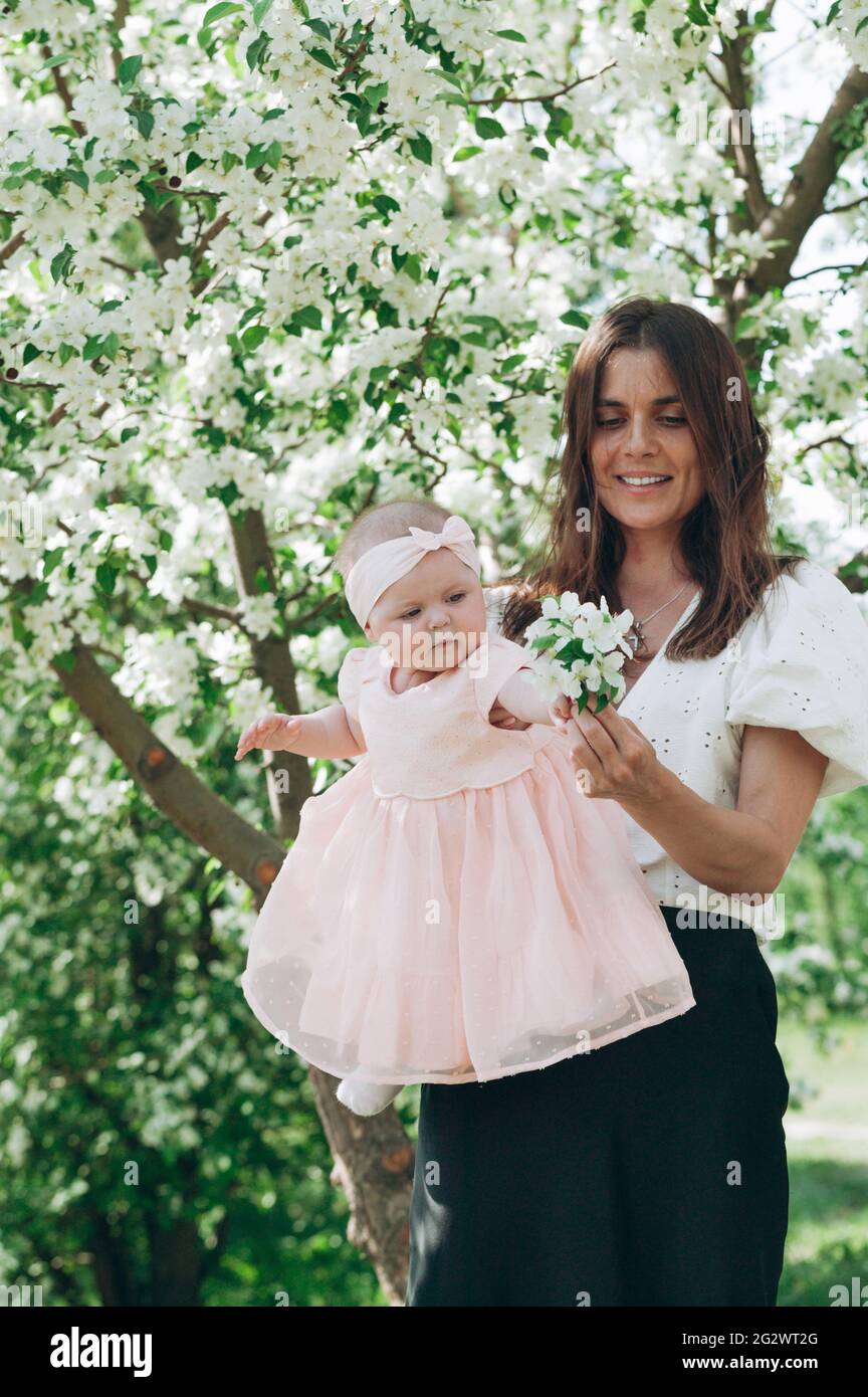 A cute beautiful young happy mom in a white shirt is holding her six month old daughter in a pink dress. Happy motherhood and happy childhood. Photo o Stock Photo