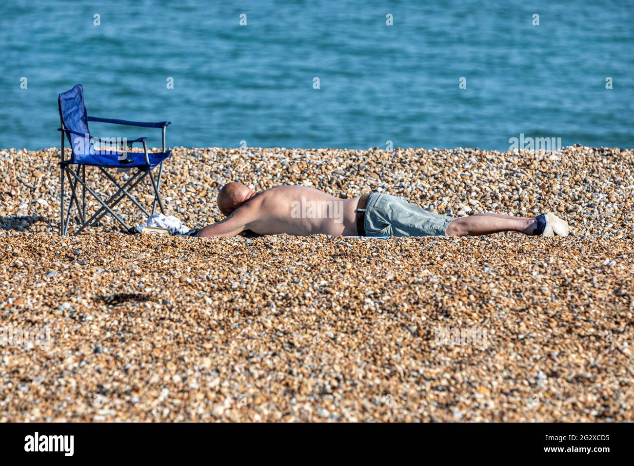 A man sunbathing on a shingle beach with a calm blue sea. Stock Photo