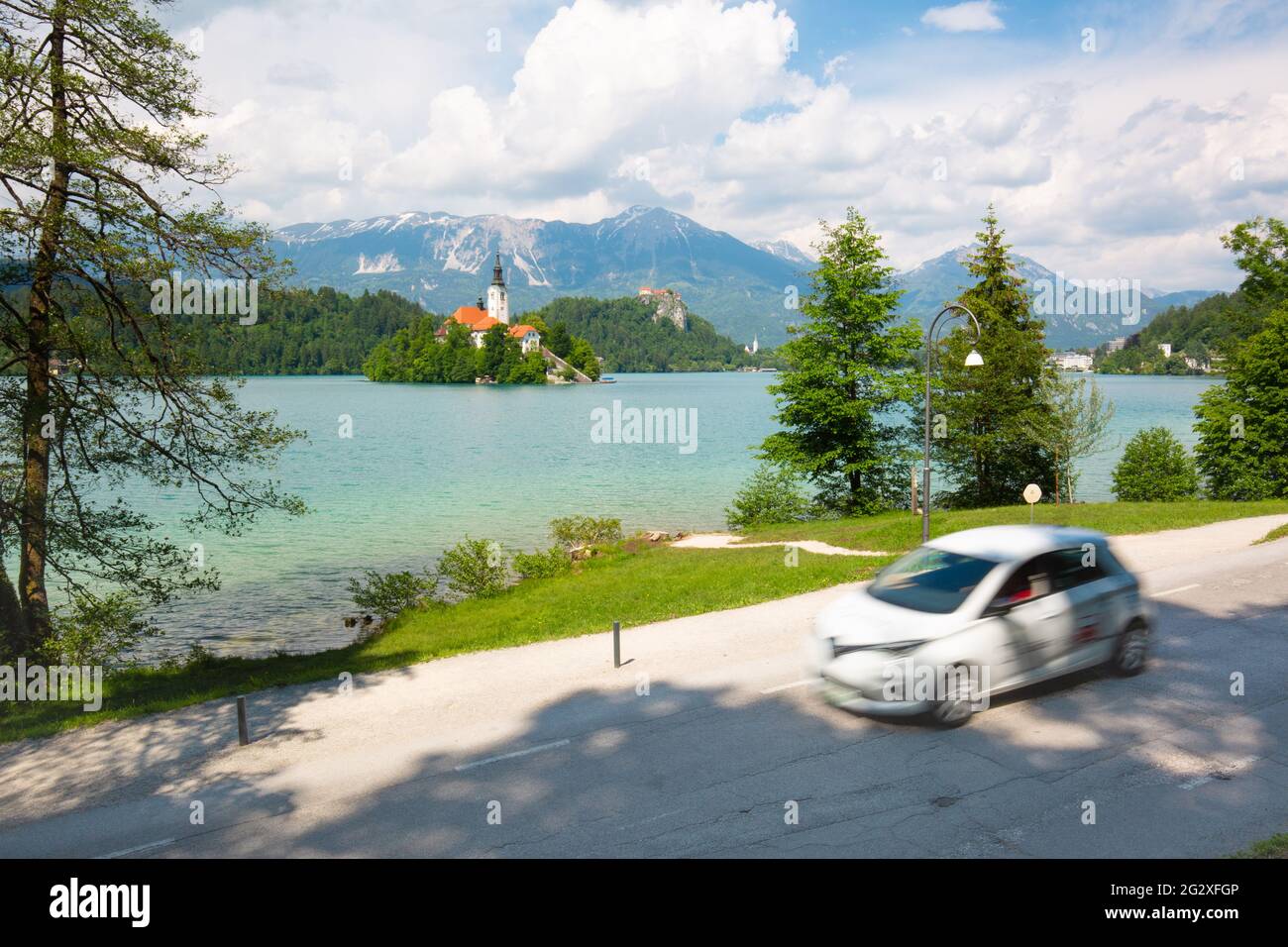 Motion blured white electric car in front of Bled lake, country of Slovenia. Travel, tourism, explore, rent a car concept. Stock Photo