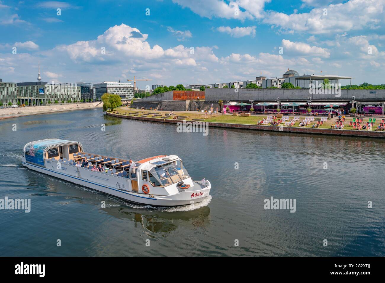 View over relaxing people at the cafe Capital Beach at Spree channel, Spreebogen, with a tour boat in historical and business downtown in Berlin, Germ Stock Photo