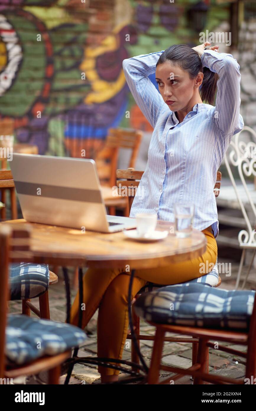 young adult caucasian female fixing her hair, sitting in the outdoor cafe with a laptop on the table and cup of coffee. remote work concept Stock Photo