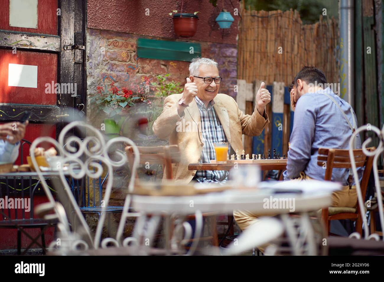 older caucasian male smiling with thumbs up. young guy with his hand over his eyes. sitting in outdoor cafe, playing chess. winning and loosing concep Stock Photo