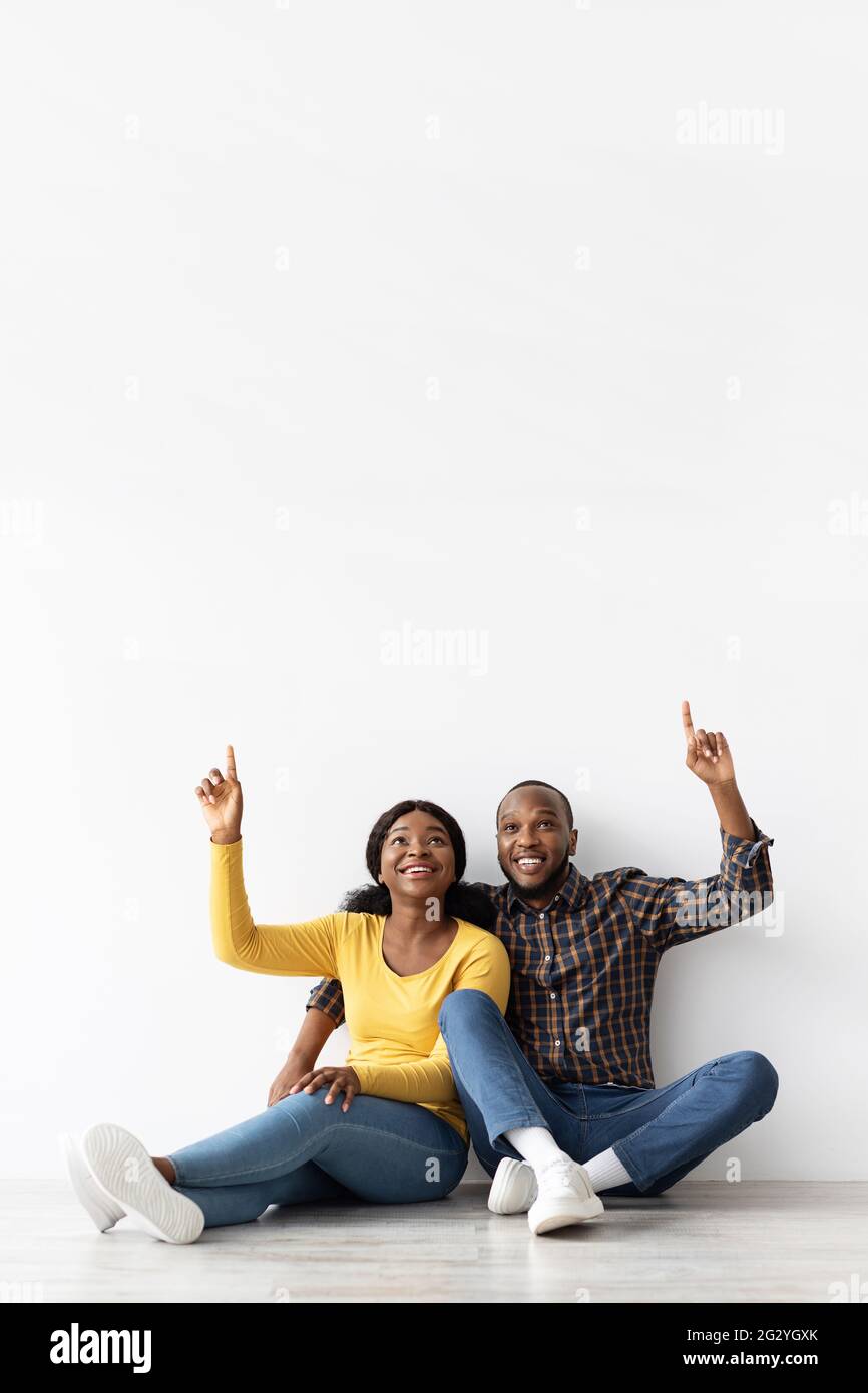 Happy African Couple Sitting On Floor Near White Wall And Pointing Up ...