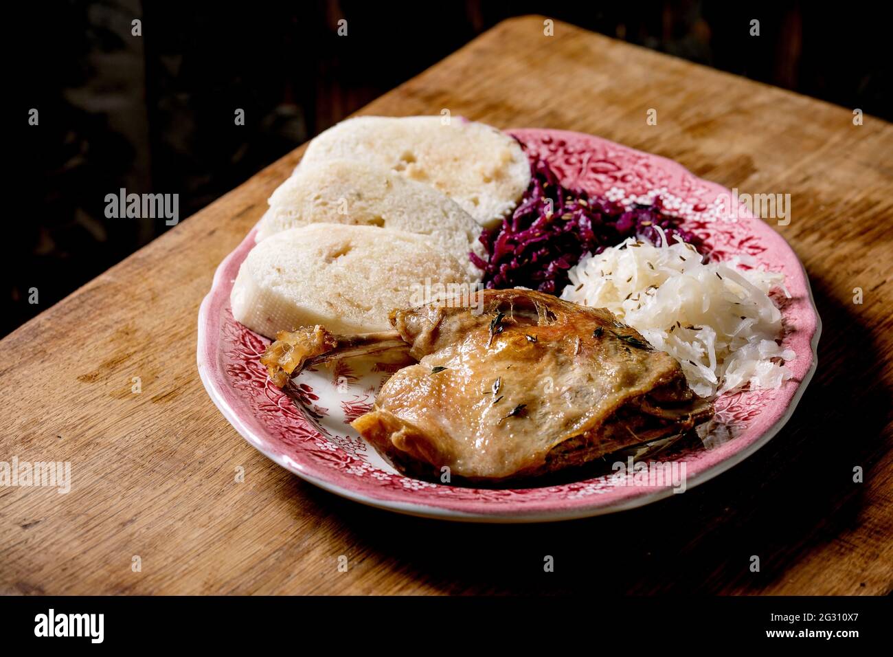 Baked duck legs with sliced boiled bread knedliks and sauerkraut in ceramic plate over brown wooden table. Traditional Czech, German and european cuis Stock Photo