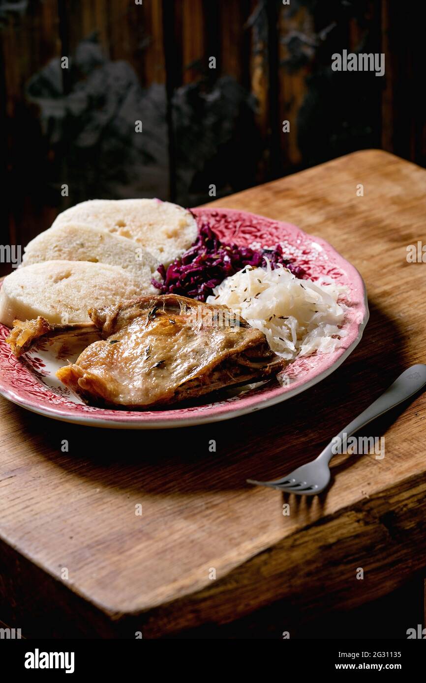 Baked duck legs with sliced boiled bread knedliks, red and white sauerkraut in ceramic plate over brown wooden table. Traditional Czech, German and eu Stock Photo