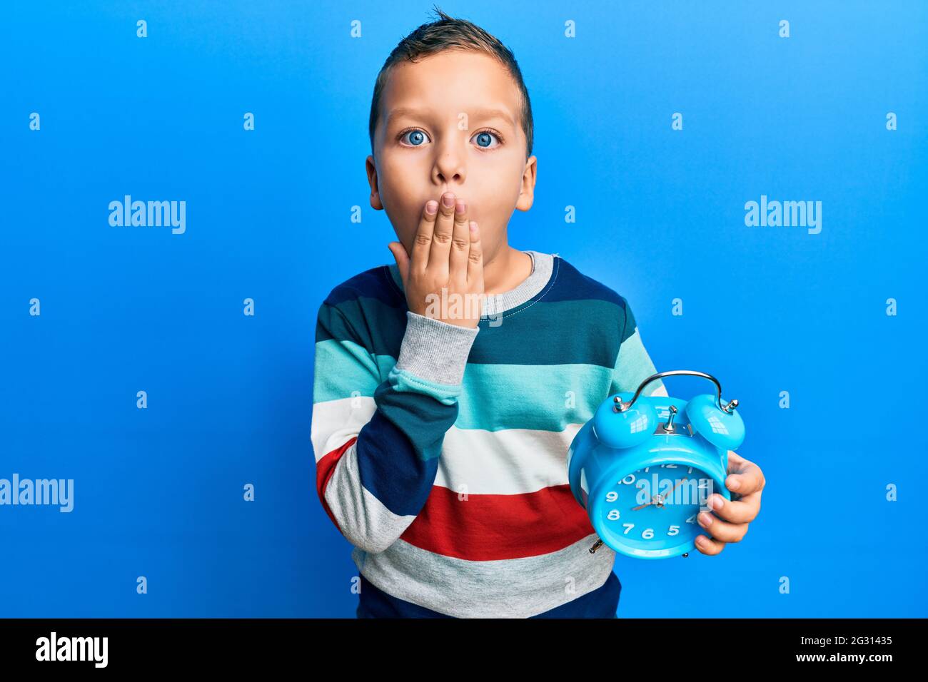 Little kid boy holding alarm clock covering mouth with hand, shocked