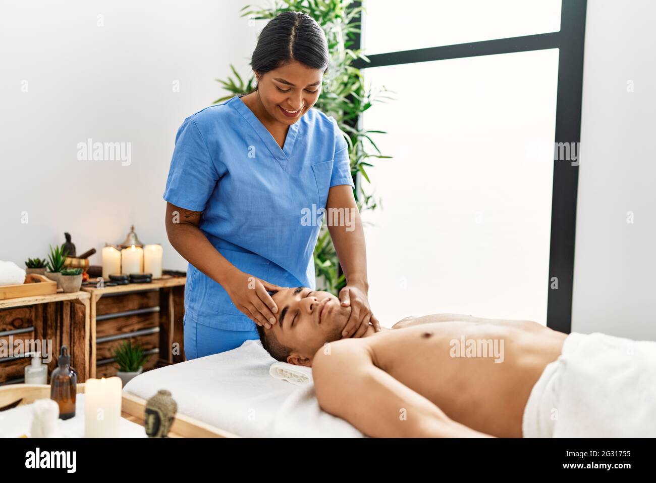 Young latin massage therapist woman giving relaxing treatment on man head at beauty center. Stock Photo