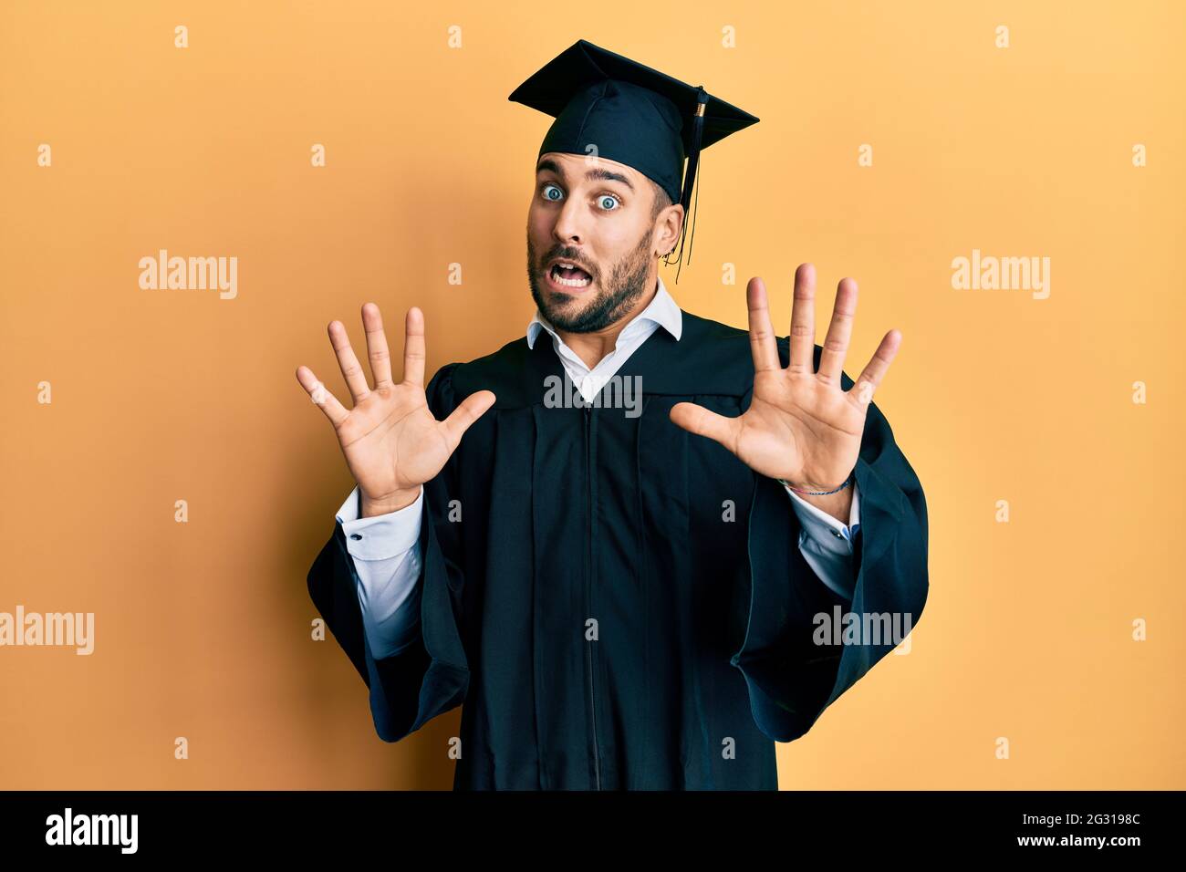 Young hispanic man wearing graduation cap and ceremony robe afraid and ...