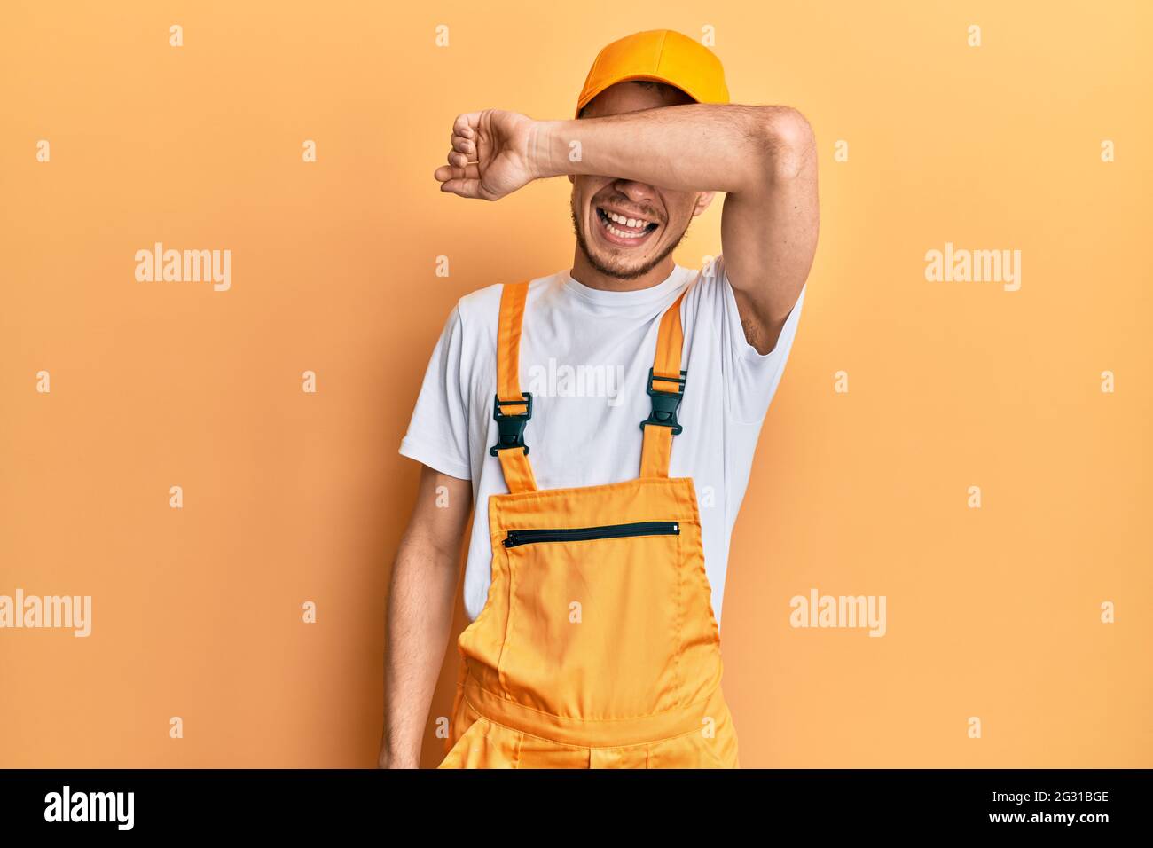 Hispanic young man wearing handyman uniform covering eyes with arm ...
