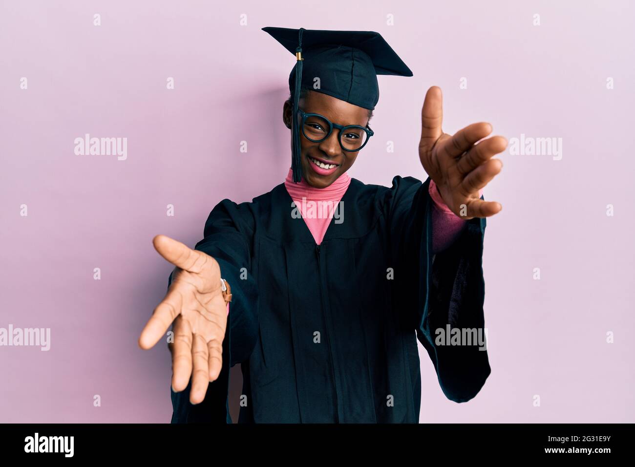 Young african american girl wearing graduation cap and ceremony robe looking at the camera smiling with open arms for hug. cheerful expression embraci Stock Photo