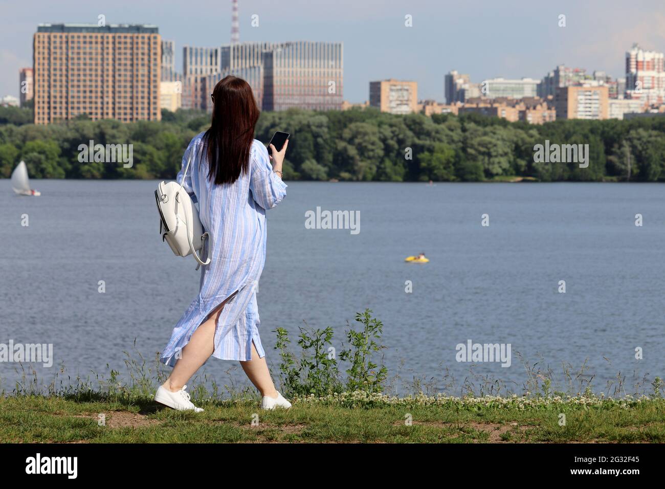 Girl in dress walking with smartphone on river and summer city background. Travel and holidays on nature Stock Photo