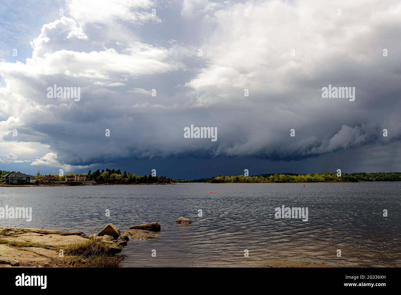 A large dark storm beneath dense clouds in the distance. It is dark blue gray under the clouds. Bright sky above.Water in the foreground. Stock Photo