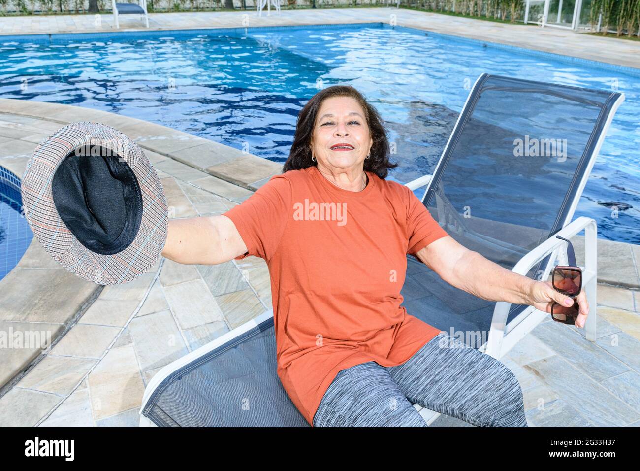 74-year-old elderly woman standing by the pool, hat in hand, happy with her retirement. Stock Photo