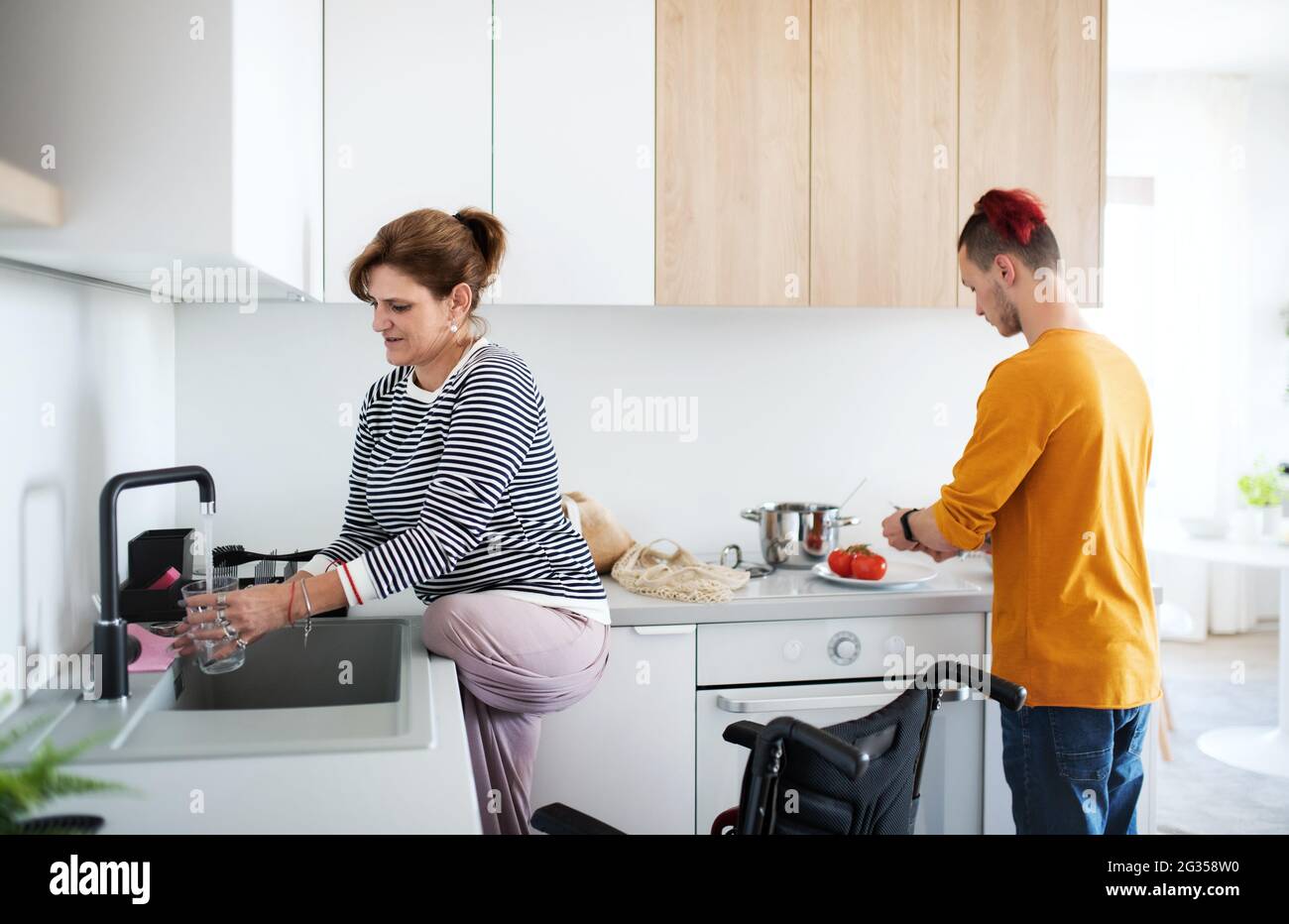 Disabled mature woman in wheelchair talking to son in kitchen indoors at home. Stock Photo