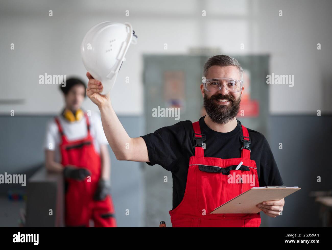 Portrait of cheerful worker with helmet and protective glasses indoors in factory looking at camera. Stock Photo