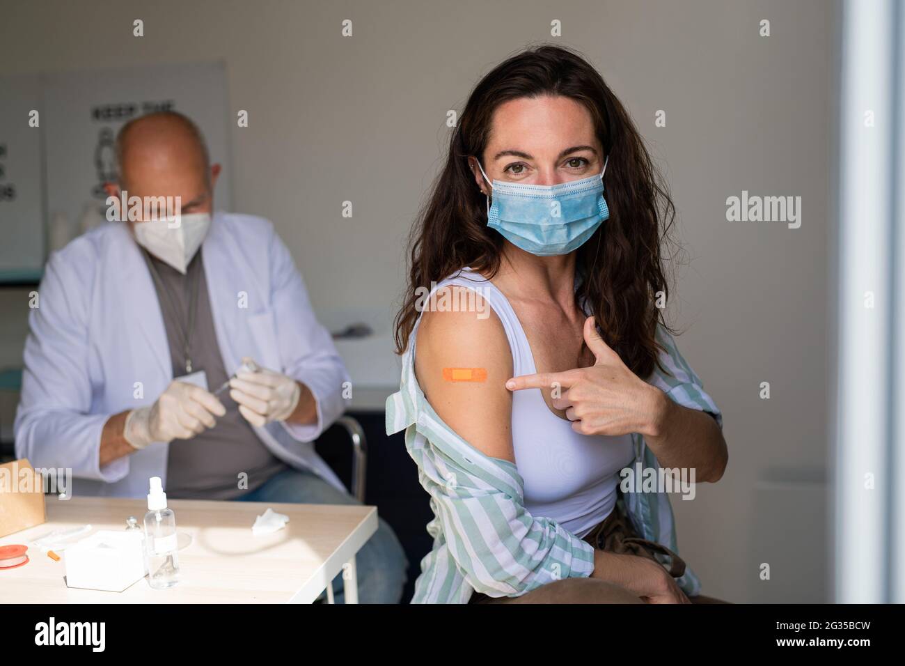 Happy young woman after covid-9 vaccination, pointing to plaster on arm and looking at camera. Stock Photo
