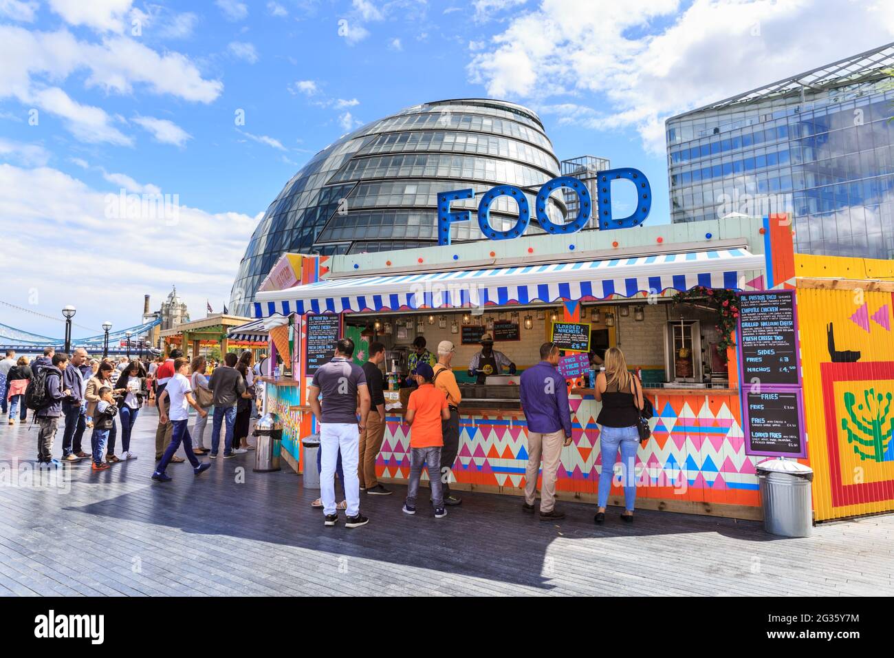People buy food from a outdoor pop up stalls at More London in the summer sun with Tower Bridge and City Hall in the background, London, England Stock Photo