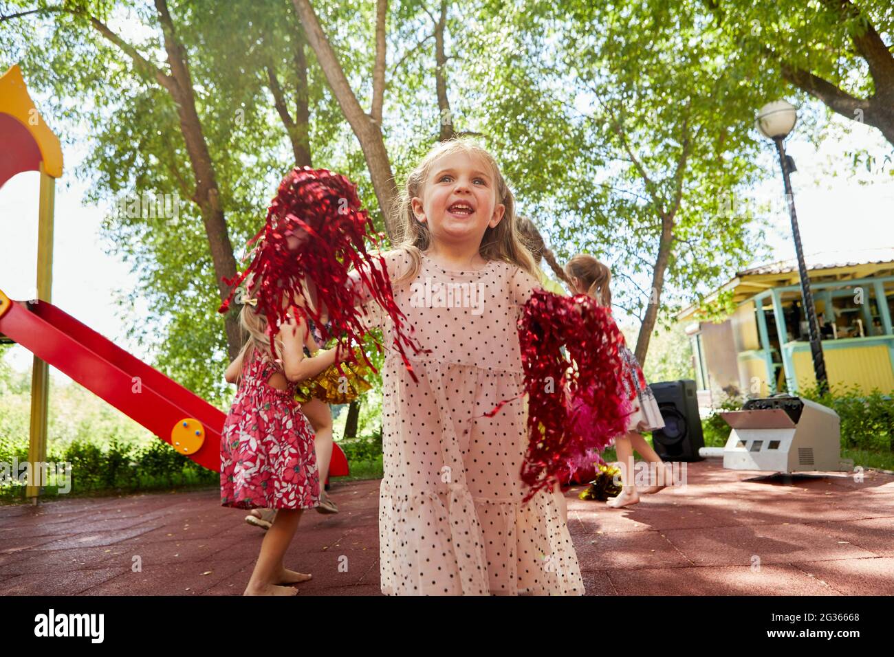Low angle of cheerful little girl with pom poms playing on playground in summer Stock Photo