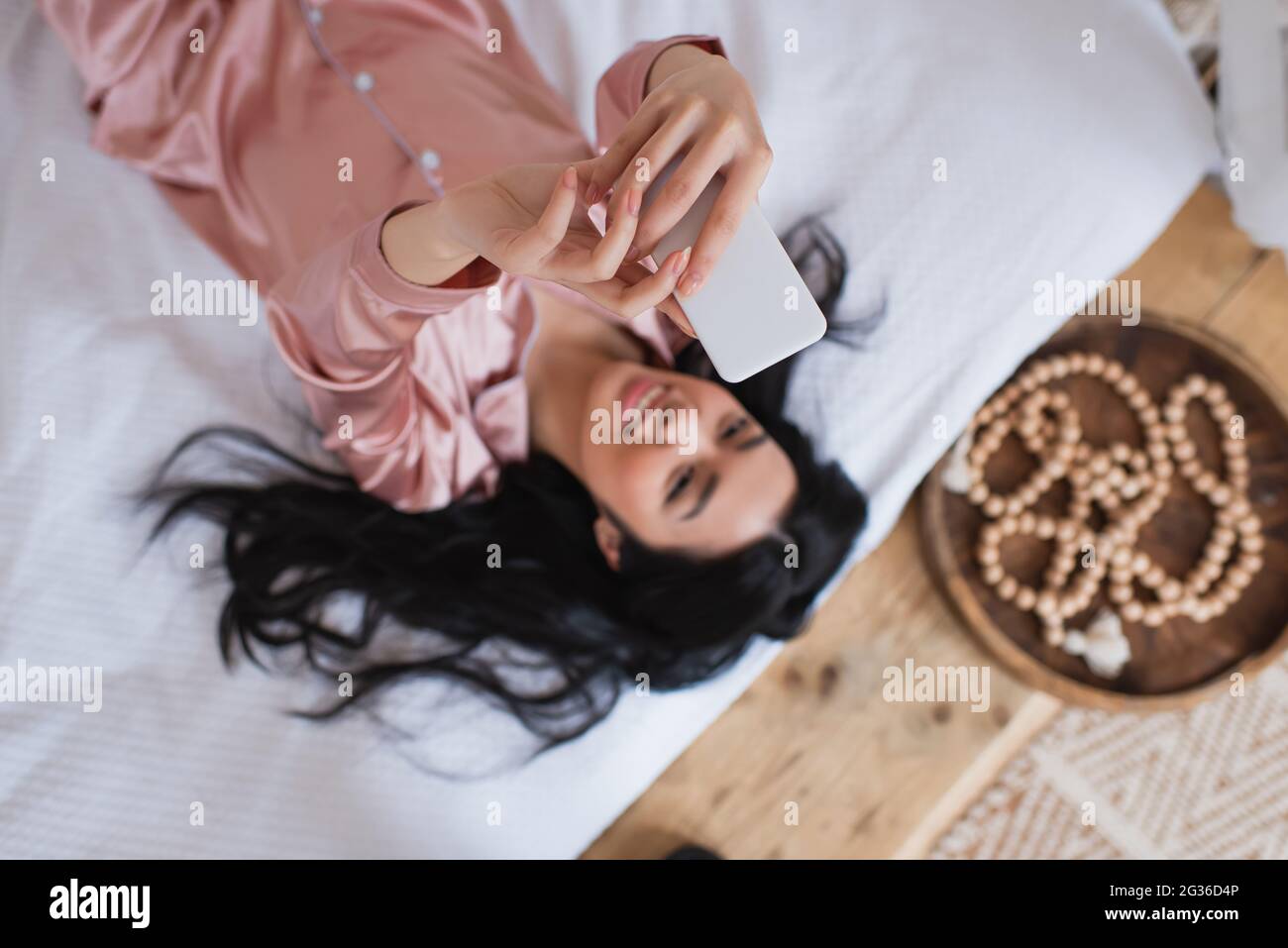 top view of smiling young asian woman in silk pajamas lying on bed and ...