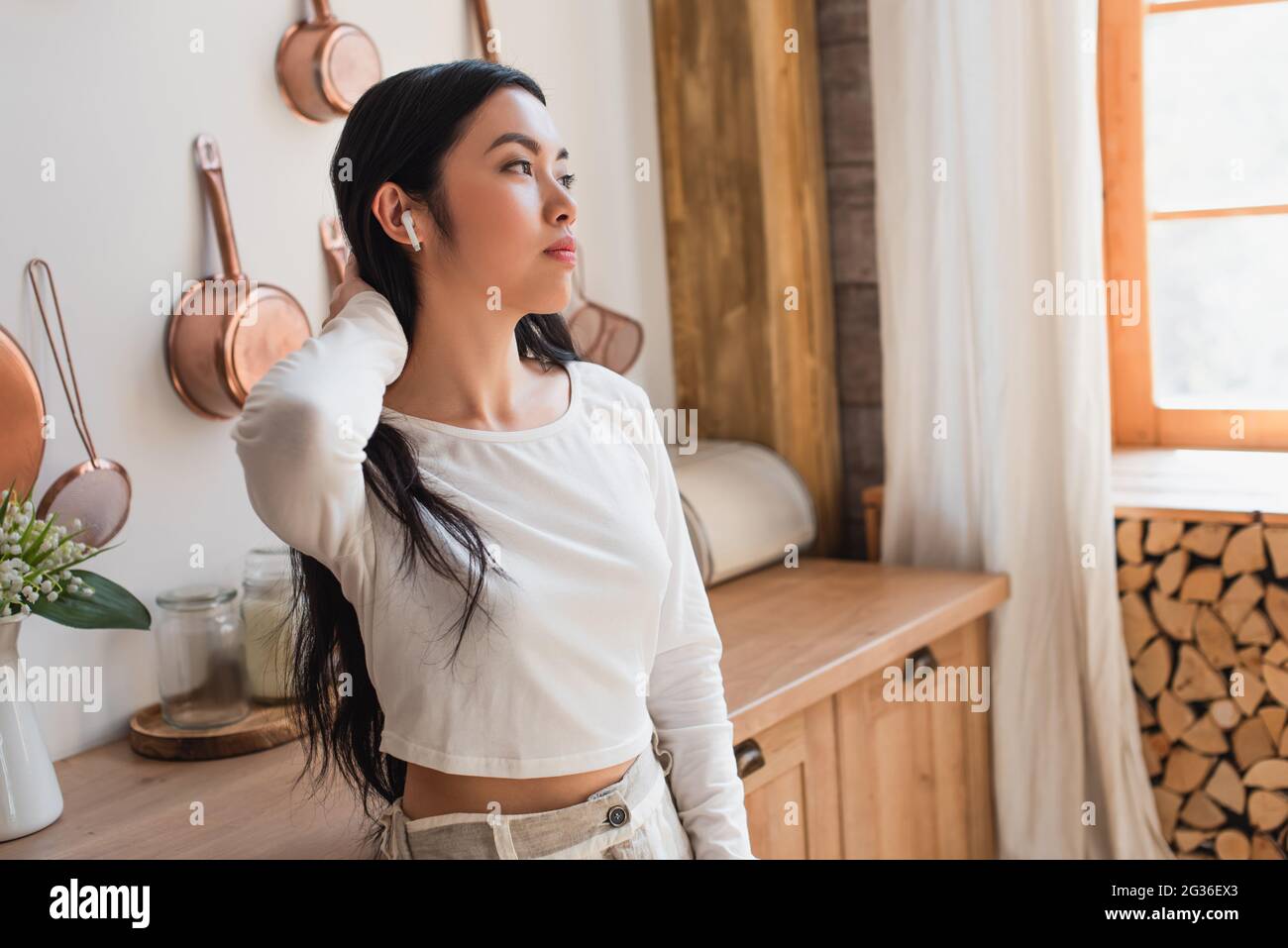 tired young asian woman in white blouse standing with earphones and holding hand near neck in kitchen Stock Photo