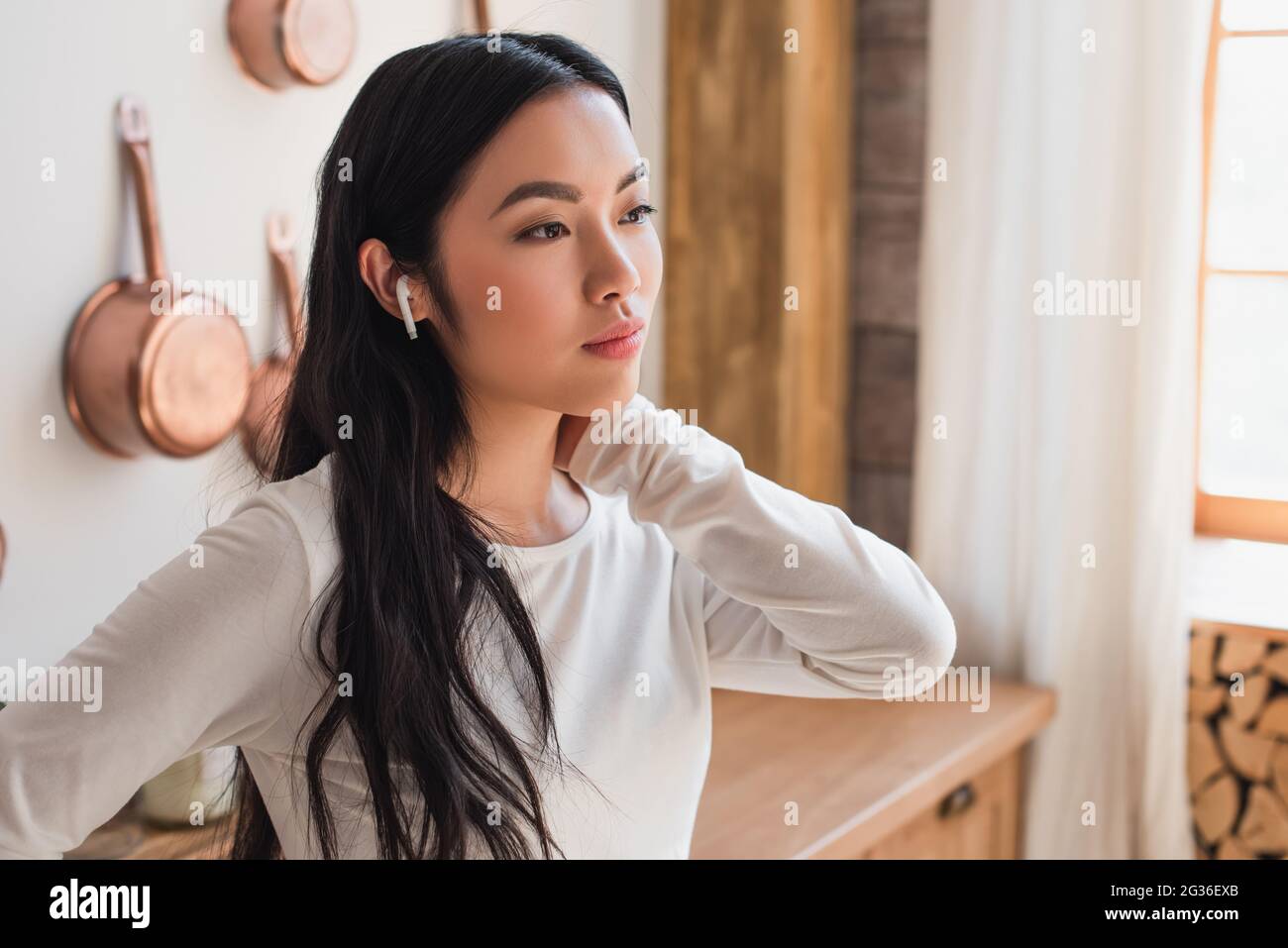 tired young asian woman in white blouse standing with earphones and holding hand near neck in kitchen Stock Photo