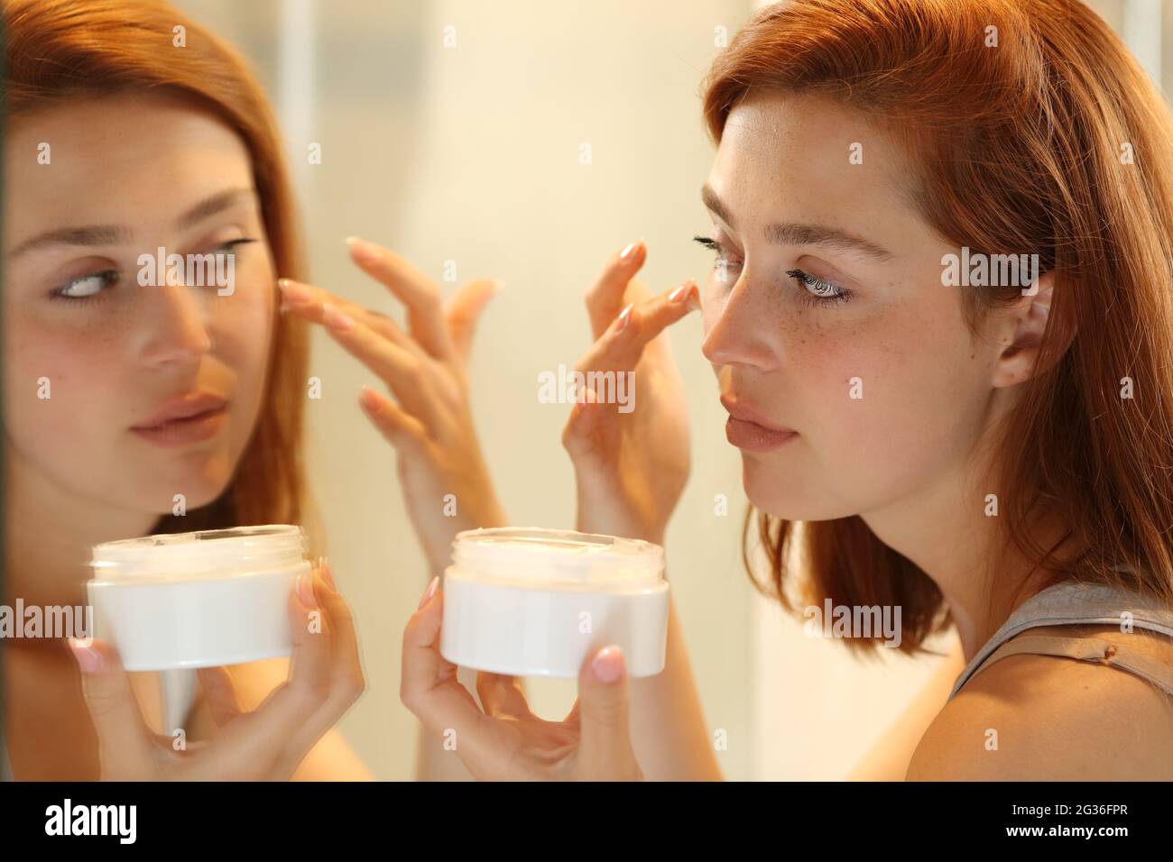 Woman hydrating face skin in front a mirror applying moisturizer cream on cheek Stock Photo