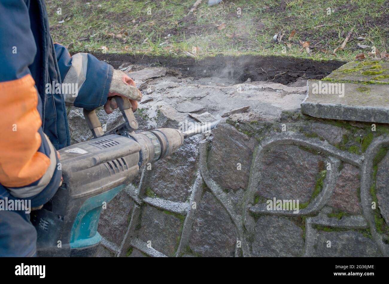 Working with rotary hammer to break off thin concrete plates of curbs, gloved hands of man dressed in overalls hold jackhammer by both handles, dust a Stock Photo