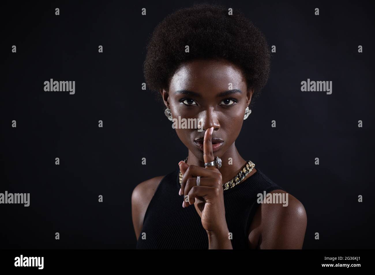 Closeup of beautiful young african american dark-skinned woman with finger on her lips showing shhh silence gesture on black background. Stock Photo