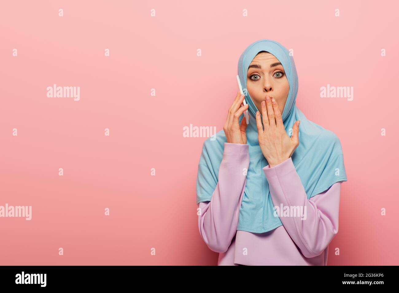 shocked arabian woman in blue hijab talking on cellphone on pink ...