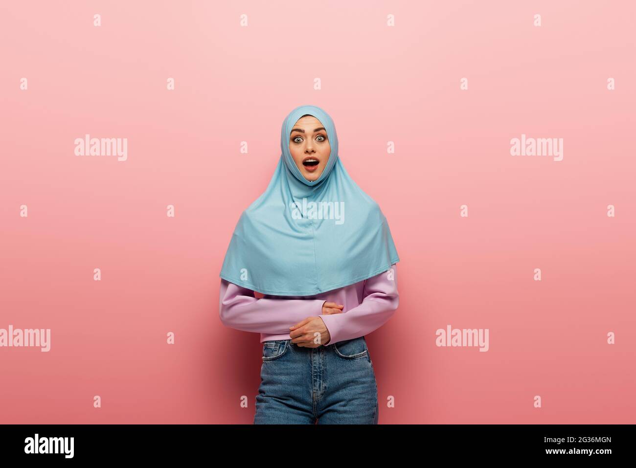 astonished arabian woman in hijab and jeans looking at camera on pink ...