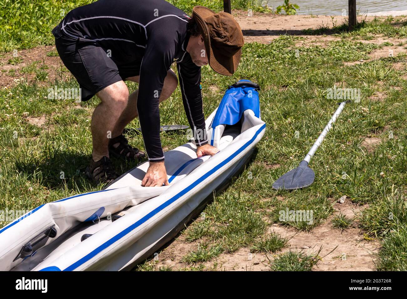 Middle aged man in brown hat, black swim shirt and shorts, folding up deflated inflatable kayak. Preparing it for storage. Paddle on ground next to it. Stock Photo