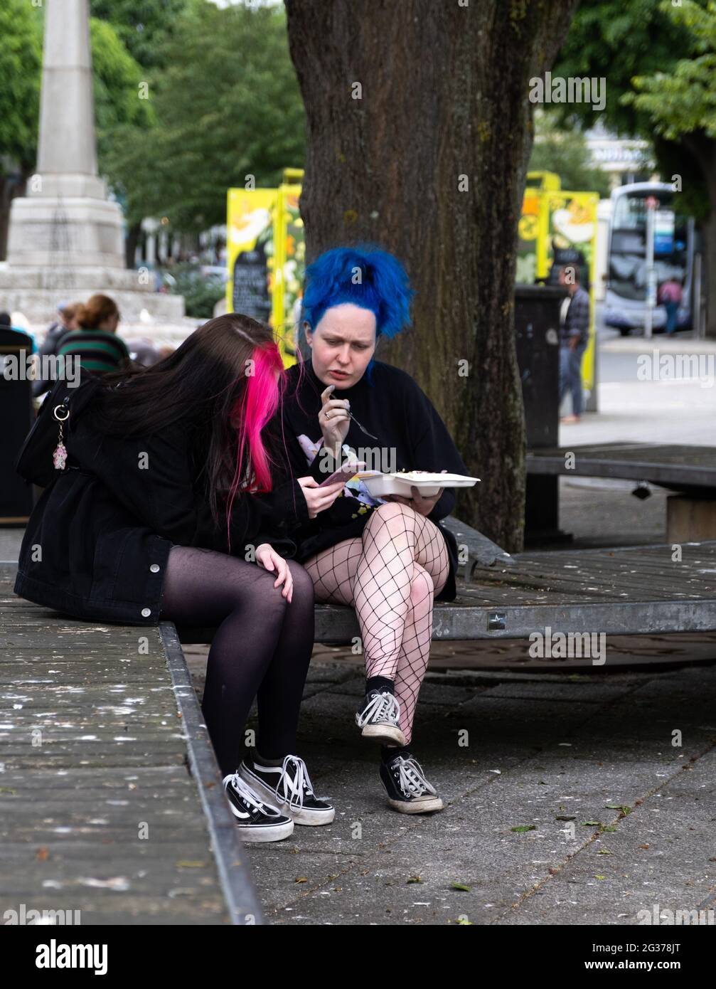 Two young girl protesters with blue and pink hair, short skirts and patterned tights sitting together in central Falmouth having a chat during G7. Stock Photo