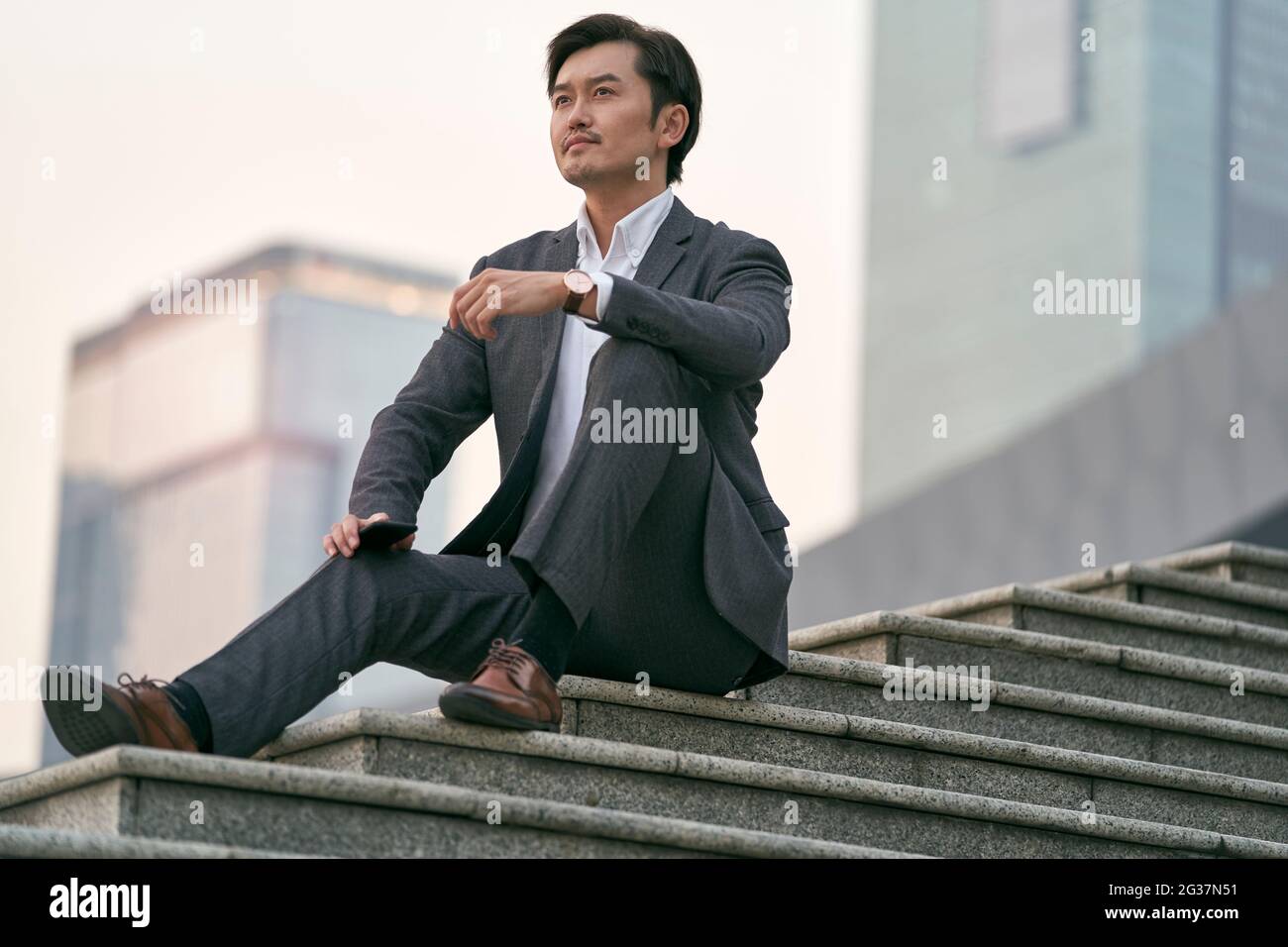 outdoor portrait of a successful asian business man sitting on steps in full suit Stock Photo