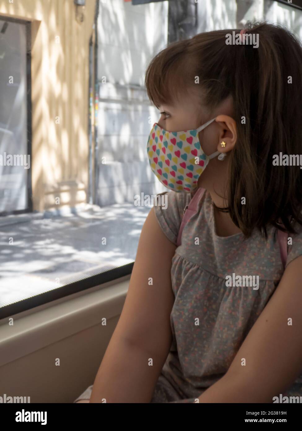 Little girl with a colorful face mask looking out the window of a bus in the city Stock Photo