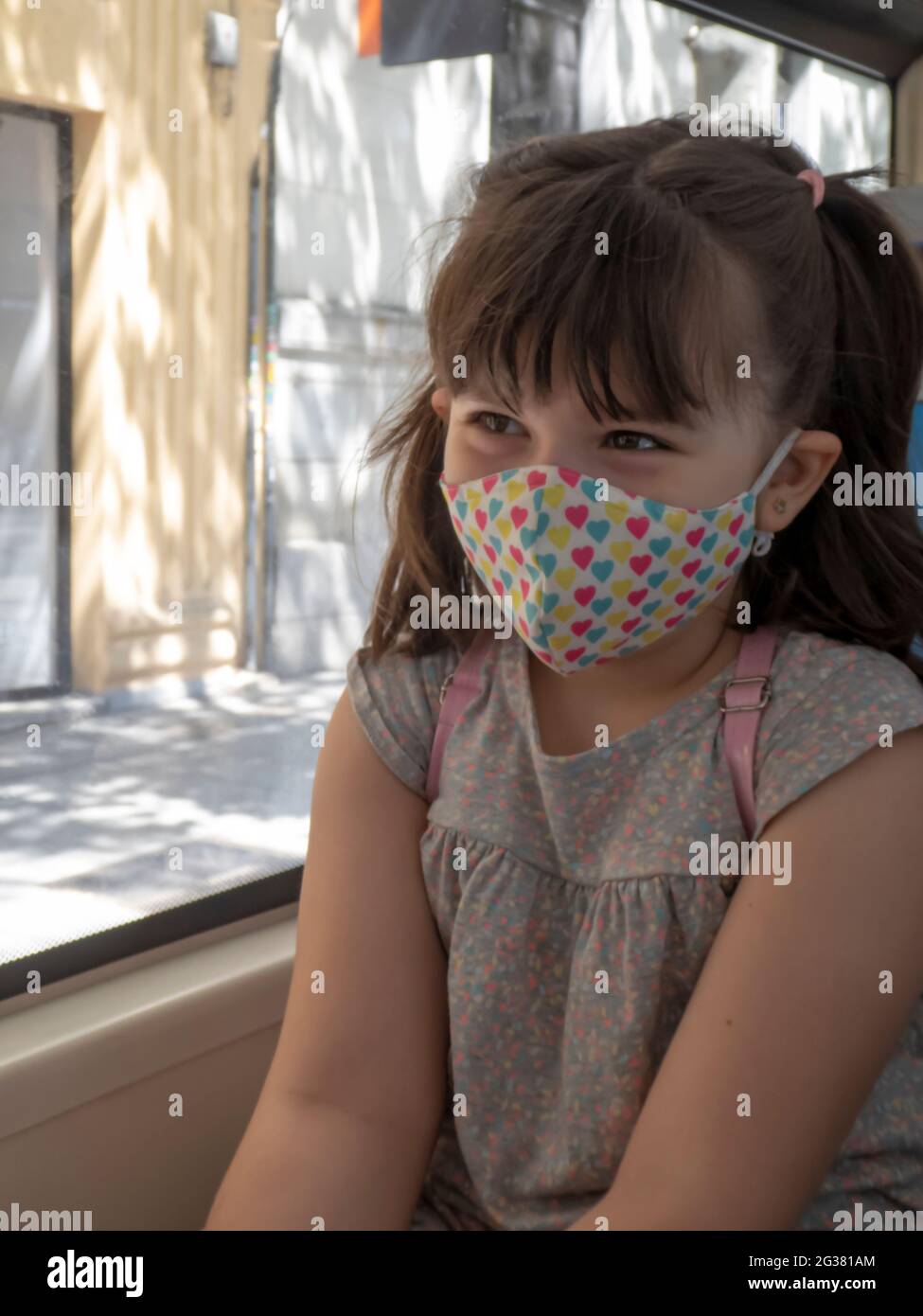 Little girl with a colorful face mask looking out the window of a bus in the city Stock Photo