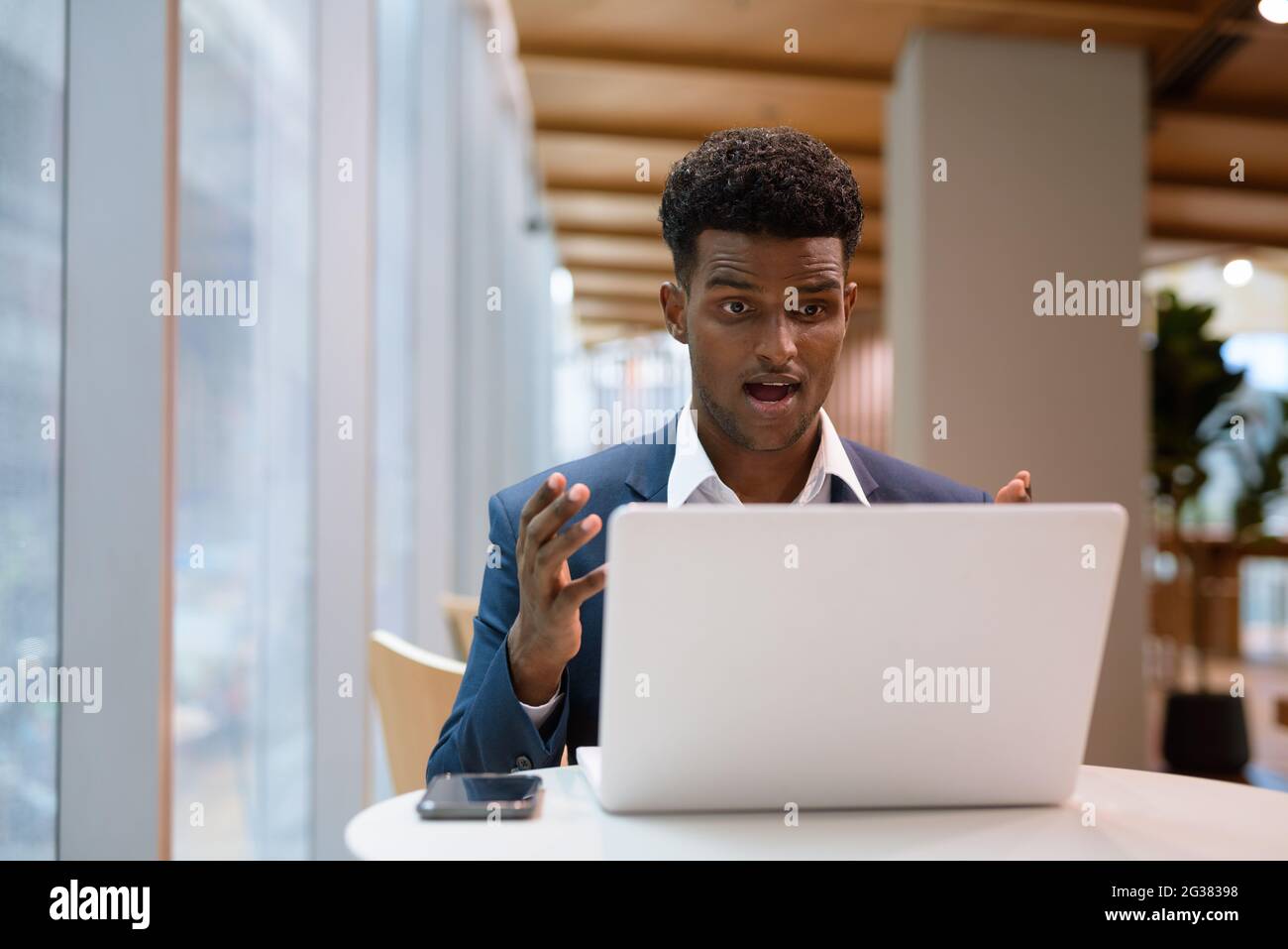 Portrait of African businessman using laptop computer in coffee shop while looking shocked and surprised horizontal shot Stock Photo