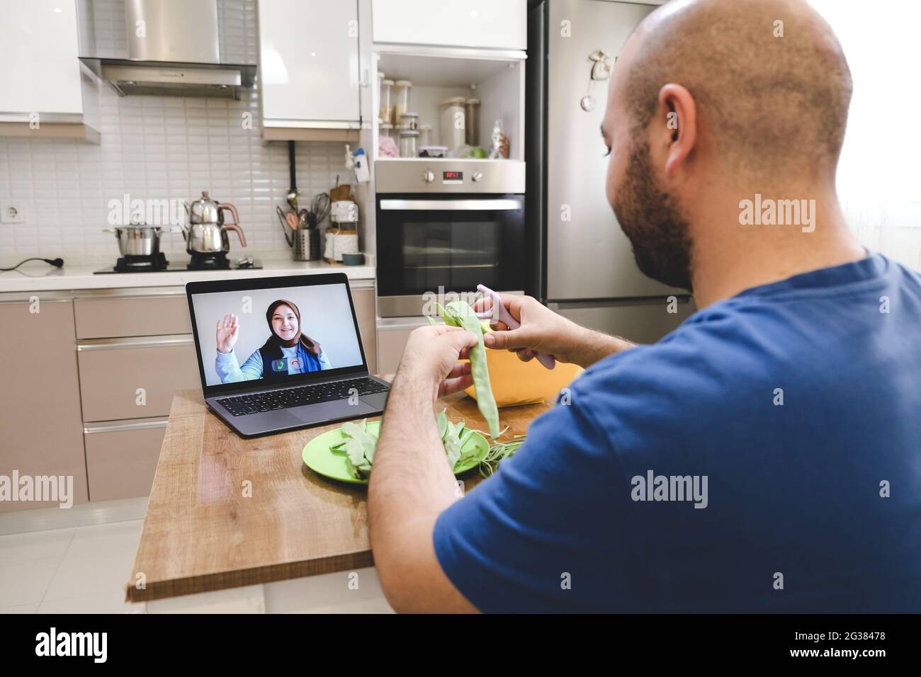 Man chatting with online video while preparing food in kitchen. Man and woman communicating via videoconference while cooking, not neglecting their wo Stock Photo
