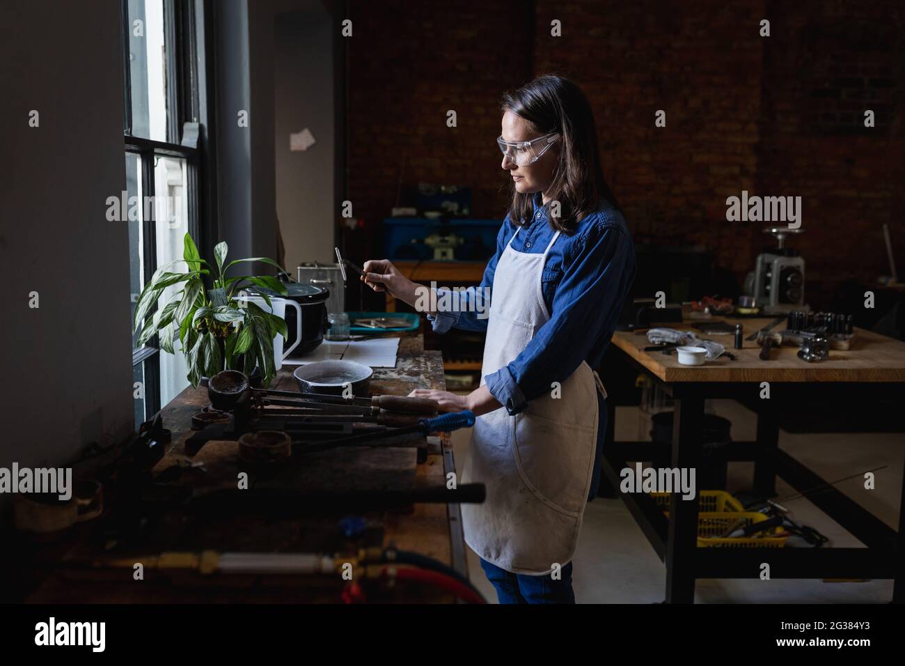 Caucasian female jeweller wearing apron and glasses, preparing materials for jewelry Stock Photo