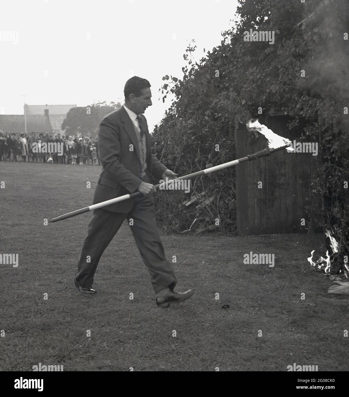 1960s, historical, evening time at a park and a man in a suit & tie holding a fire-lighter, a long pole lighted at one end with a flame, around the entrance to a large public bonfire covered with tree brances to set the the structure alight, Fife, Scotland, UK. Stock Photo