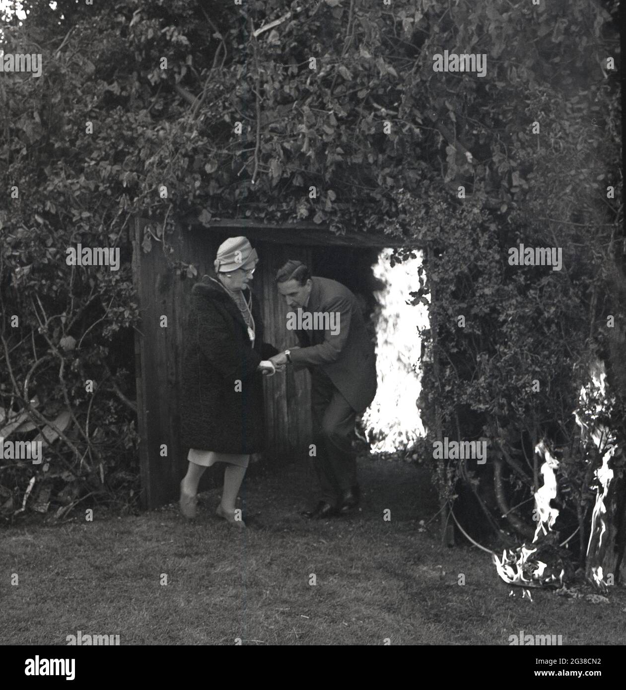 1960s, historical, early evening time at a park and an elderly lady helping a gentleman at a entrance to a large public bonfire covered with tree brances which he has just lit. Stock Photo