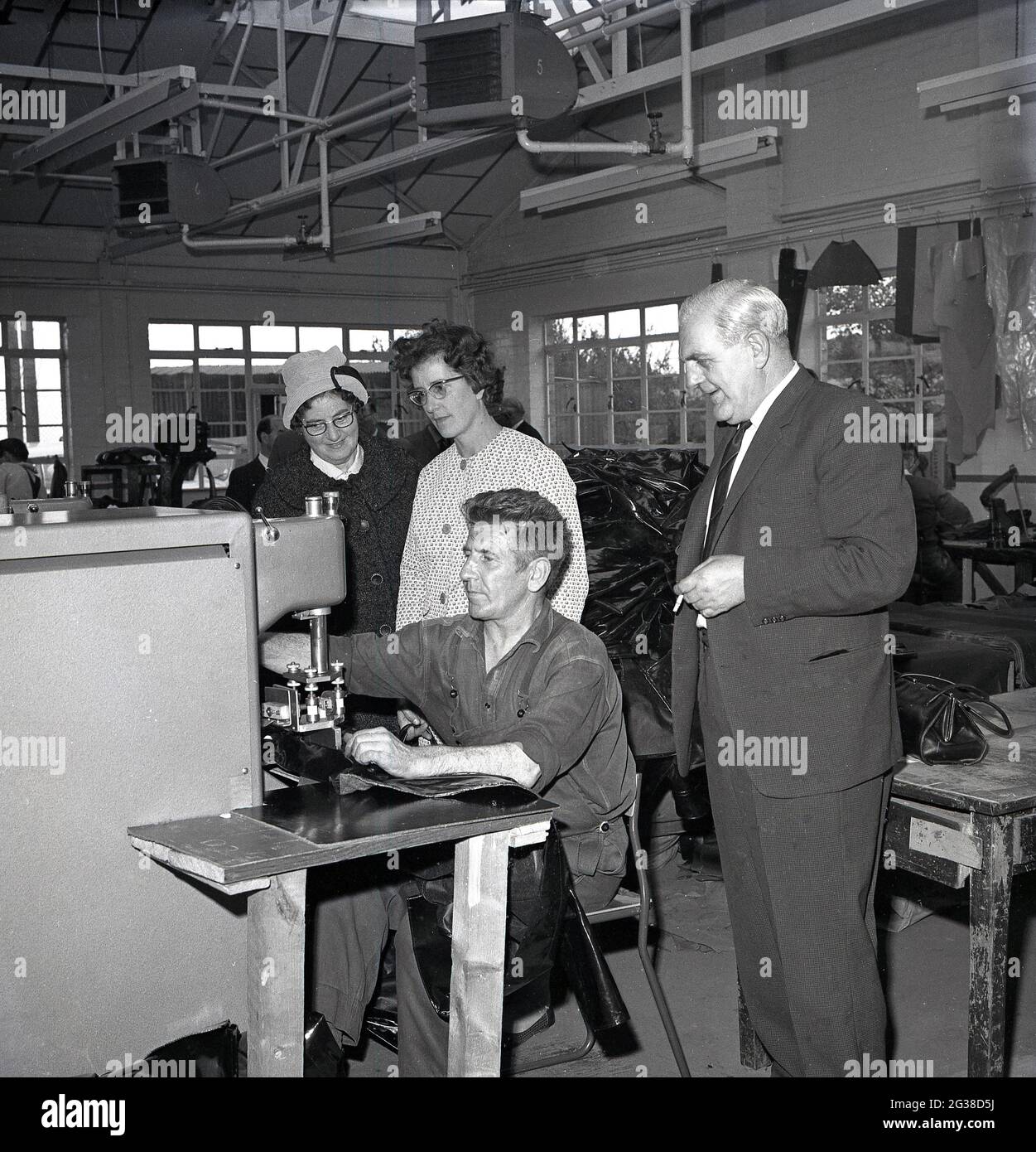 1960s, historical, inside an indistrial unit, two lady local councilors and a company Director, standing by a worker, a  former mineworker being re-employed in industry, using a large machine to stitch or seal leather goods such as bags together, Cowdenbeath, Fife, Scotland. Stock Photo