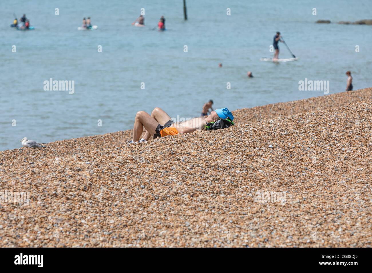 A man sunbathing on a shingle beach with people enjoyng watersports in the distance. Stock Photo