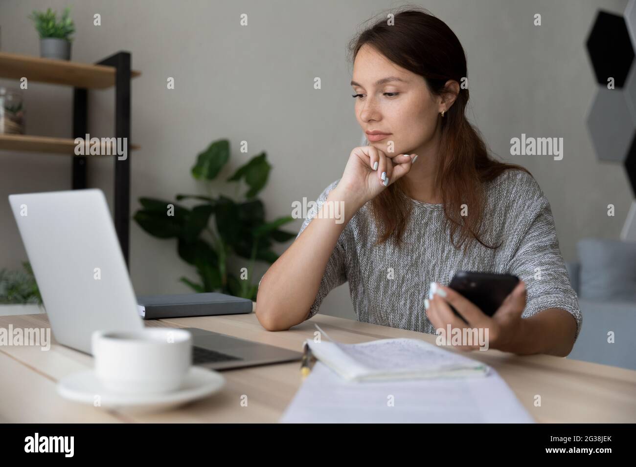 Thoughtful confident businesswoman looking at laptop screen, holding smartphone Stock Photo
