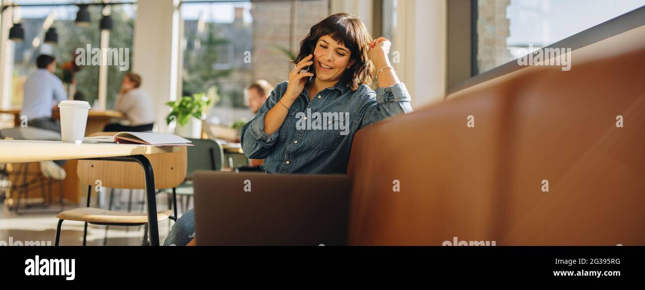 Businesswoman sitting on sofa in coworking space and talking on cell phone. Woman looking at laptop and speaking on mobile phone. Stock Photo