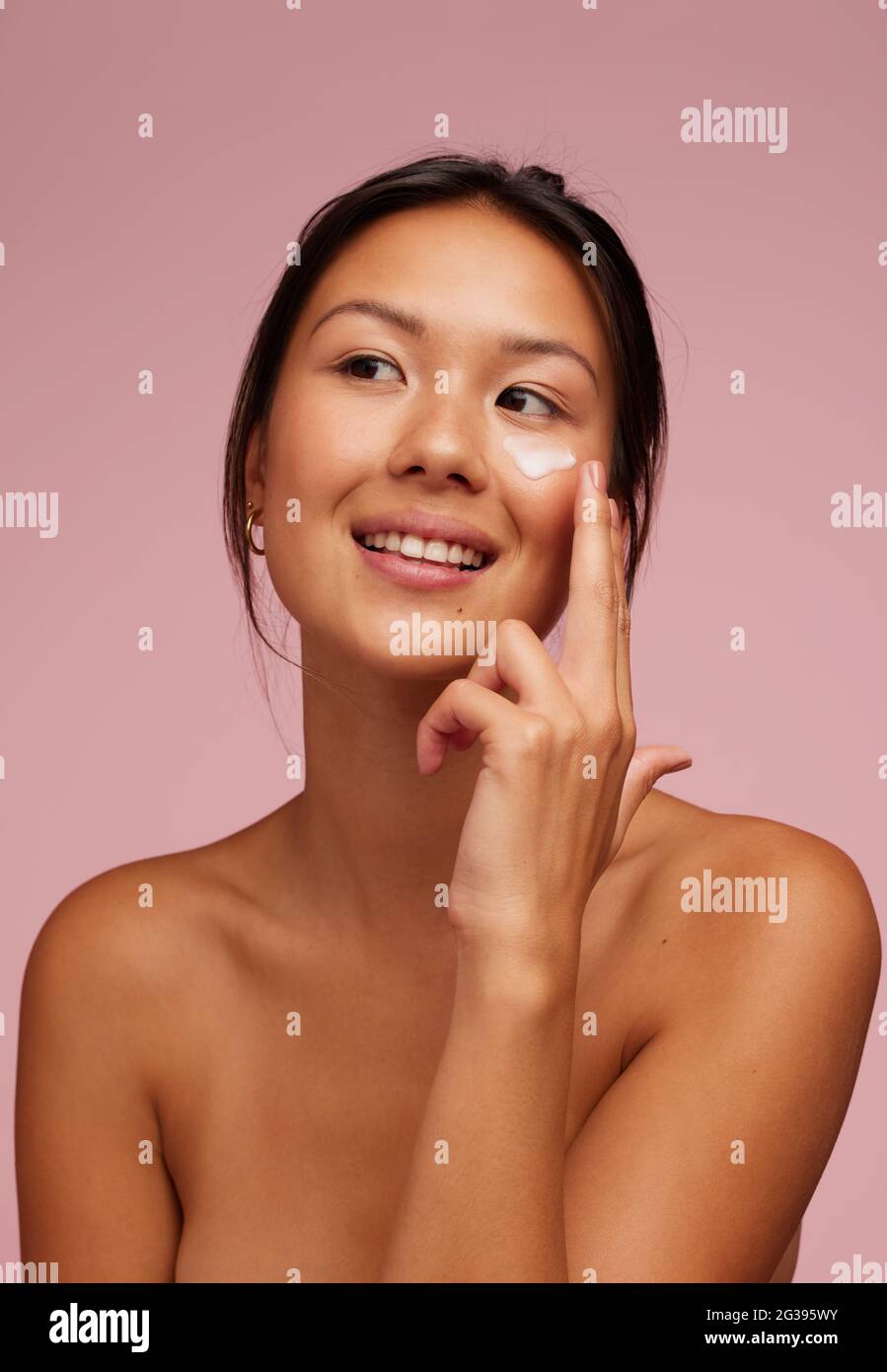 Portrait of a beautiful woman applying moisturizer cream on her face. Woman using beauty product on her facial skin and looking away. Stock Photo