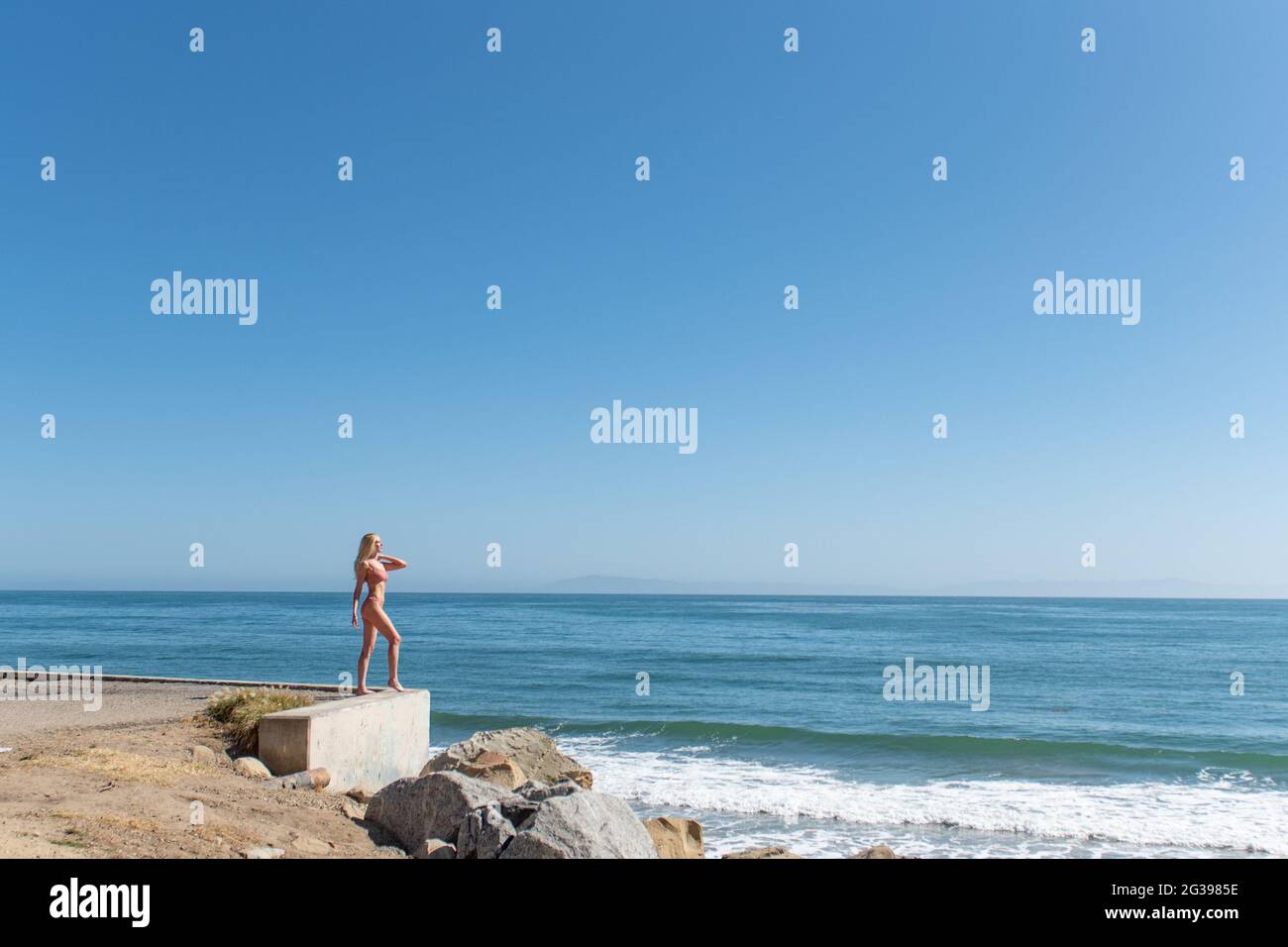 Beautiful blond hair female model wearing two piece binki swimsuit while looking out into the large Pacific Ocean from the rocky shoreline. Stock Photo