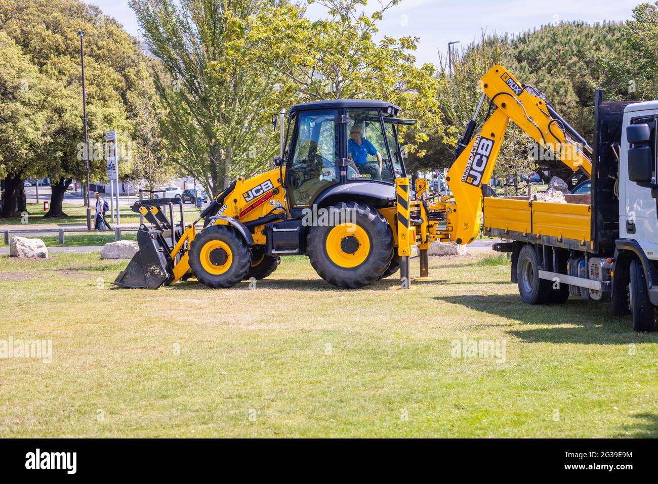 A yellow JCB 3CX Eco backhoe loader working in a park in Southsea, Portsmouth, Hampshire, south coast England with its stabilisers set down Stock Photo