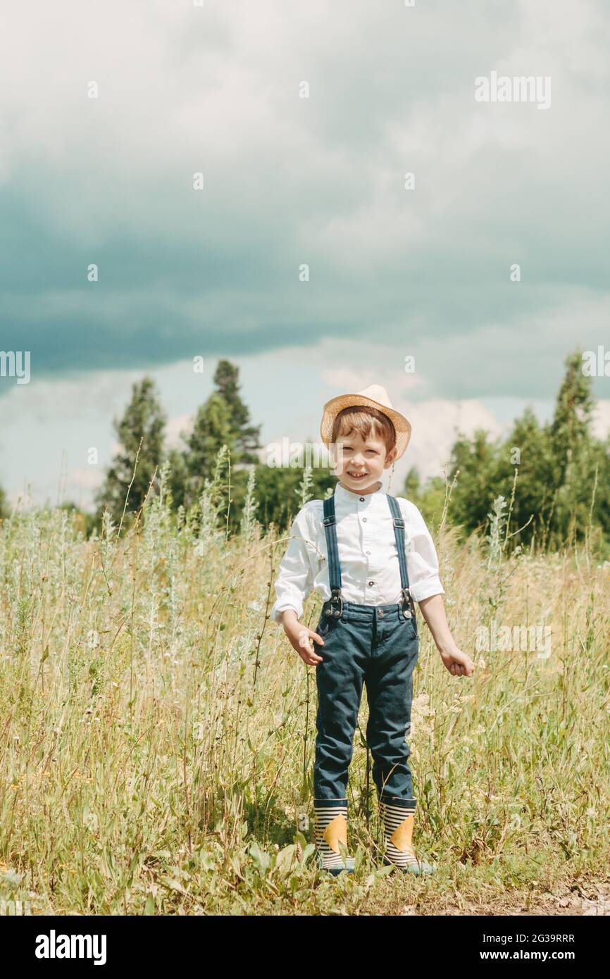 Little farmer on a summer field, cute little boy in a straw hat. boy with a flower stands in a field. boy in rubber boots and a white shirt. Country s Stock Photo